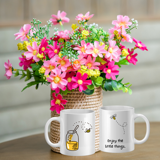 Two white mugs with bee designs and text next to a vase of colorful flowers on a wooden surface.